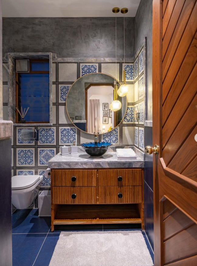 A luxurious bathroom interior design by Olangana in Bangalore, featuring vibrant blue and white patterned wall tiles, a chic wooden fluted vanity with a marble countertop, a distinctive blue vessel sink, a round gold-framed mirror, and modern spherical pendant lights. The floor features solid blue tiles, complementing the rich textures and bold color palette.