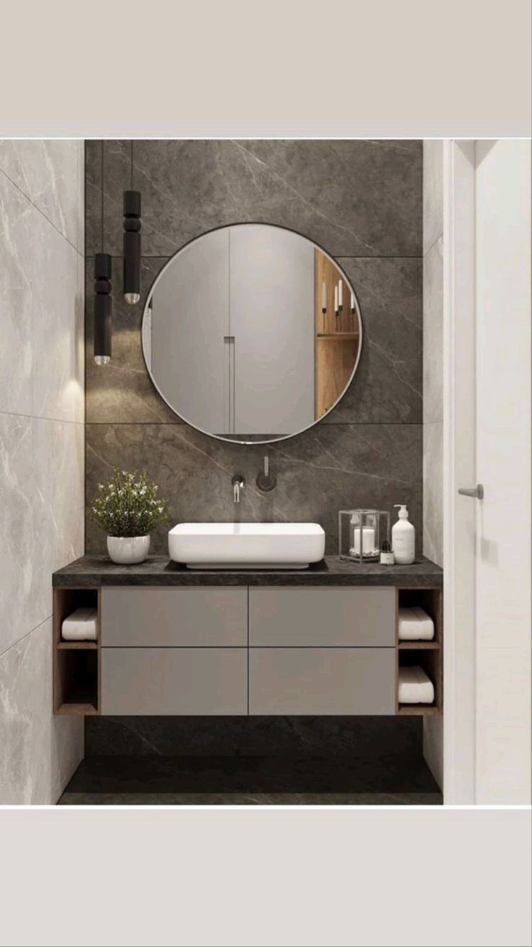 A luxurious and modern bathroom interior design by Olangana in Bangalore, featuring a floating vanity with light grey cabinets, a sleek white vessel sink, and a dark marble-effect accent wall. Two elegant black pendant lights flank a large circular mirror, enhancing the sophisticated ambiance with Olangana's signature Interior Design style.