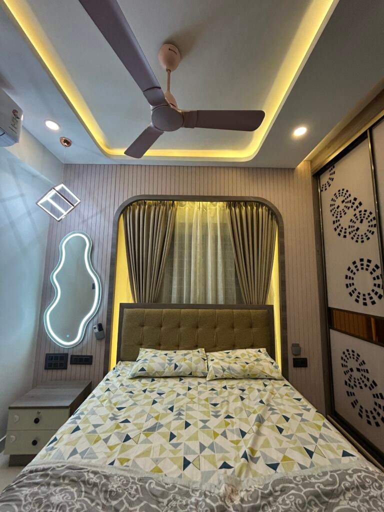 Modern master bedroom with warm lighting, sleek sliding wardrobe, POP ceiling with fan, stylish bed setup, curtains, and dressing table with mirror lighting by Olangana Design’s.