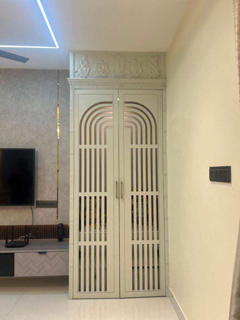 The pooja room combines modern interior design with traditional Indian religious elements. The wall behind the prayer area features sacred symbols such as Nama, Shanka, and Chakra, adding spiritual meaning to the space. The design creates a calm and divine atmosphere for daily prayers. This type of pooja room design is often included in modern Bangalore homes by Olangana Design’s.